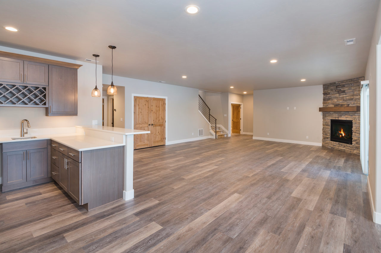 An empty basement with mini kitchen bar, hanging pendant lights, recessed lights on the ceiling, and a fireplace in one corner