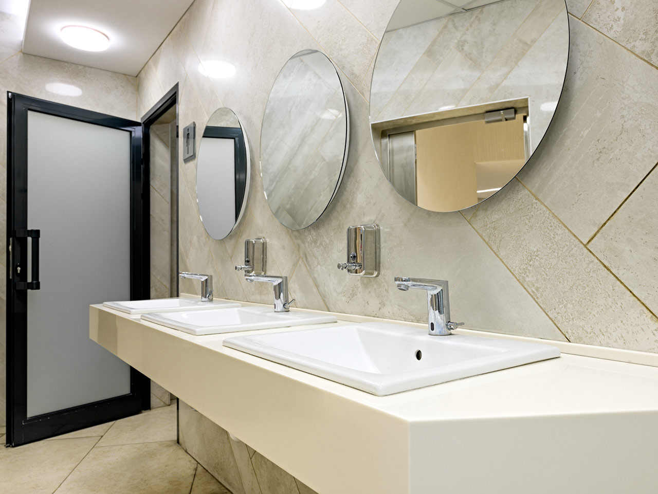A bathroom vanity area, three sinks on a countertop, and three circular mirrors against a tiled wall